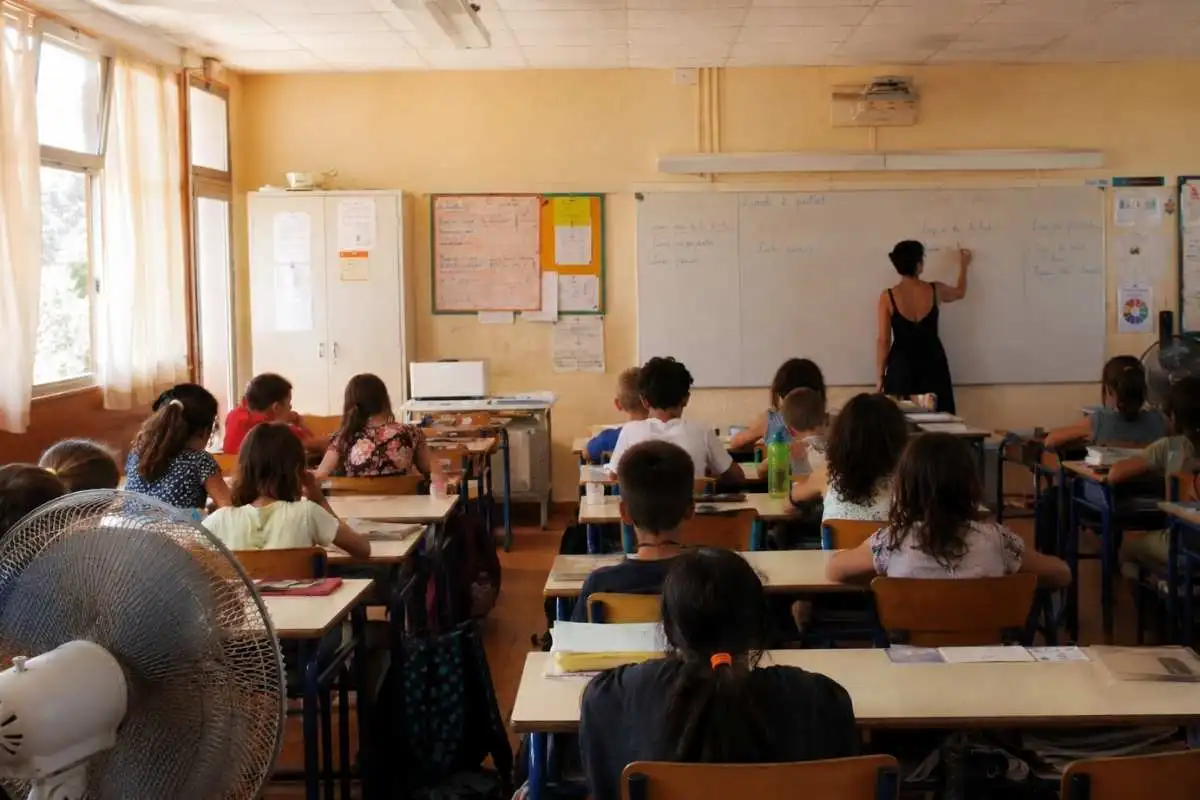 Salle de classe d'école primaire bondée et surchauffée, avec ventilateur visible, illustrant l'inconfort thermique des élèves et enseignants.